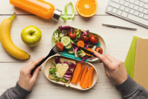 Healthy snack at office workplace. Businesswoman eating organic vegan meals from take away lunch box at wooden working table with computer keyboard